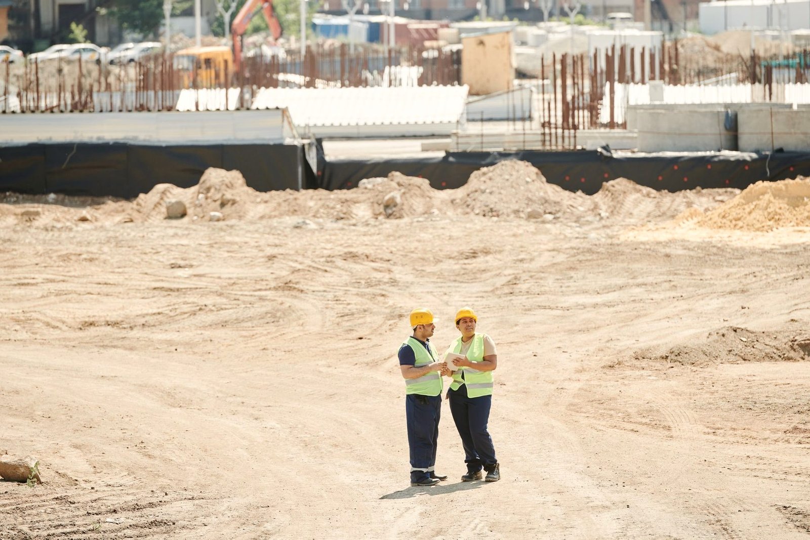 Two workers in hard hats and high visibility vests crossing a sand covered site.