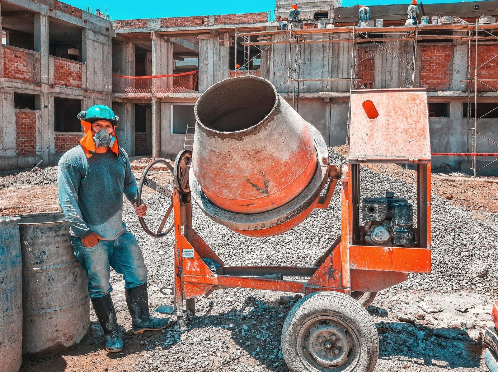 A worker operating a drum concrete mixer beside an unfinished block structure.