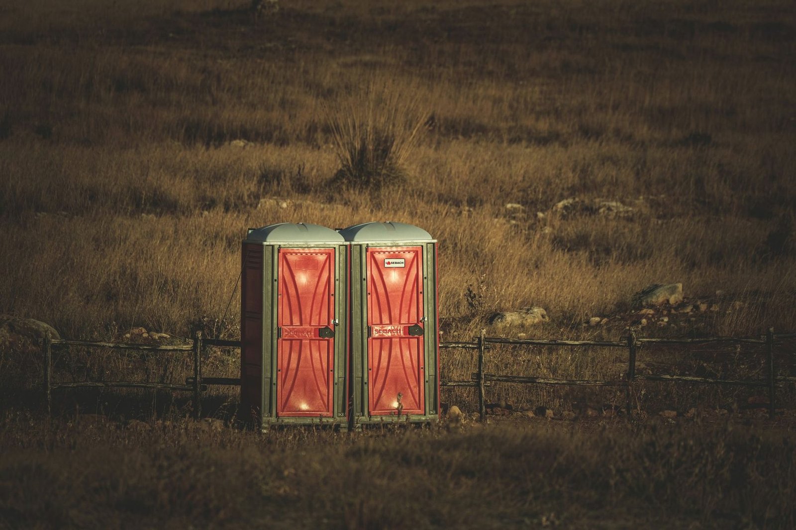 Two portable toilet units behind a post and wire fence at the edge of a dry field.